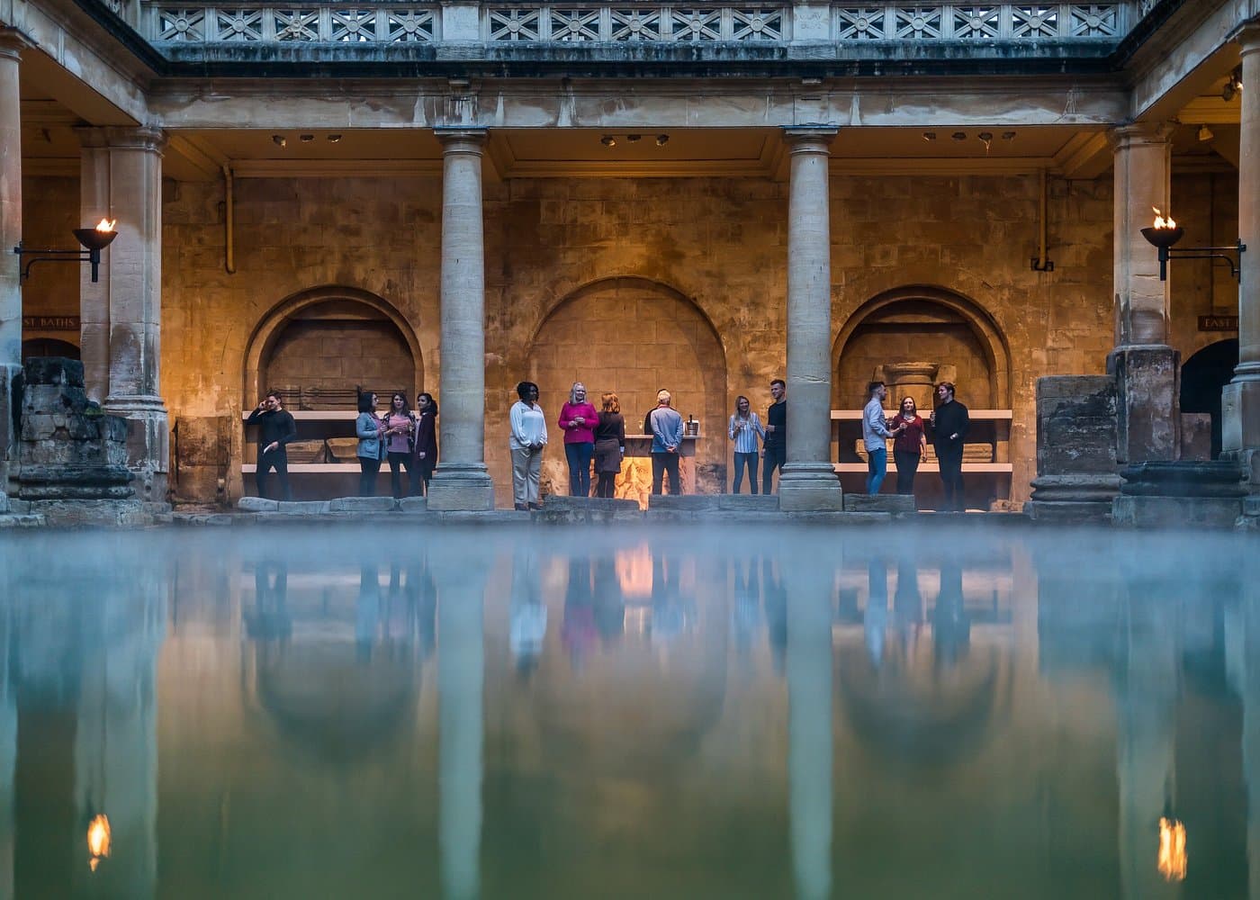 Steaming Great Bath at the Roman Baths in Bath, with visitors on the stone terrace.