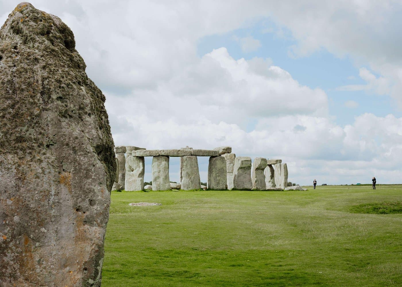 Stonehenge on a bright day, framed by a lichen-covered sarsen stone in the foreground.